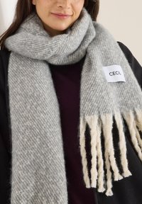 Szaro-biały szalik w wzory, wykonany z miękkiego materiału, z skręconymi frędzlami na końcach oraz prostokątną metką z logo "CECIL".