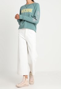 Turkusowa bluza z logo "Abercrombie" w kolorze żółtym, białe szerokie spodnie i jasnoróżowe wysokie trampki. Materiał wydaje się miękki.