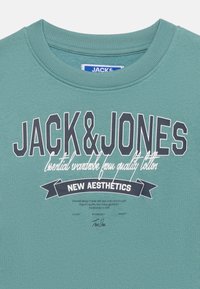 Τυρκουάζ φούτερ με στρογγυλή λαιμόκοψη, με τη φράση "JACK & JONES" σε μεγάλα γράμματα, κείμενο με γραφικό χαρακτήρα από κάτω και πανό με το κείμενο "NEW AESTHETICS" στην πρόσοψη.