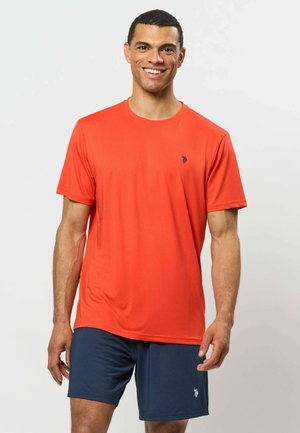 Mann lächelt, trägt ein orangefarbenes kurzärmeliges T-Shirt und marineblaue Shorts, steht vor einem schlichten hellen Hintergrund.