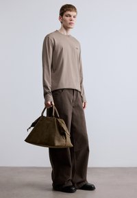 Bézs színű, hosszú ujjú ing finom logóval, laza barna nadrággal párosítva. Egy személy nagy, olívazöld bőr táskát tart. Fekete cipő.