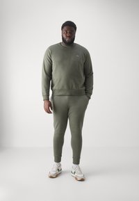 Uomo in piedi che indossa un felpa e pantaloni jogger verde oliva abbinati, sneakers bianche con dettagli verdi, contro uno sfondo chiaro e neutro.