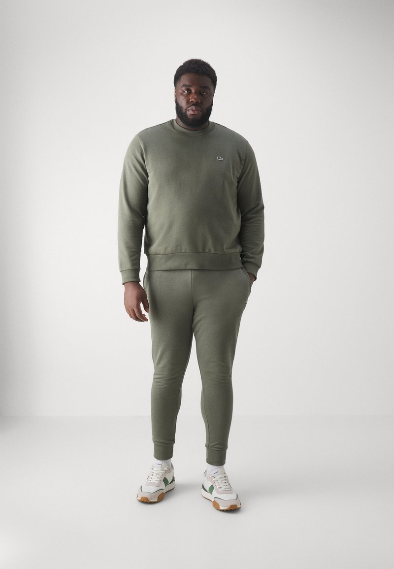 Man staat in een bijpassende olijfgroene sweatshirt en joggingbroek, witte sneakers met groene details, tegen een effen lichte achtergrond.