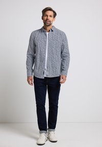 Camicia a maniche lunghe, con fantasia in blu e bianco, indossata sopra una T-shirt bianca, abbinata a jeans scuri e sneakers bianche.