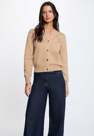 Frau trägt einen beigen, geknöpften Cardigan mit einem bestickten Wappen und dunkle, weit geschnittene Jeans und steht vor einem schlichten hellen Hintergrund.