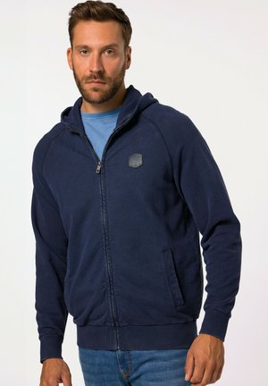 Sudadera con cremallera azul marina, hecha de tejido suave, con una cremallera en el frente, bolsillos laterales, puños acanalados y un pequeño parche con el logo en el pecho.