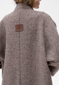 Bruin herringbone-patroon jas met een gestructureerde afwerking, met een zichtbaar merkje op de achterkant nabij de kraag.