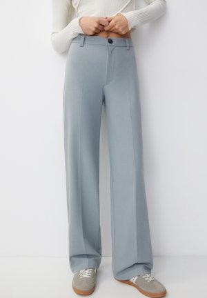 Woman wearing high-waist, light blue wide-leg trousers paired with grey lace-up sneakers, standing against a white wall.