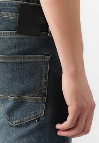 Jeans de mezclilla azul oscuro con una etiqueta de cuero negra, costuras amarillas y un bolsillo trasero con detalles curvados. Mano descansando cerca.