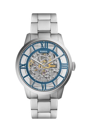 Orologio da polso Fossil in argento con quadrante trasparente che mostra gli ingranaggi meccanici, numeri romani blu e cinturino in metallo argento.