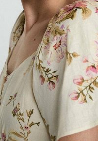 Robe florale en tissu léger avec une base crème, ornée de motifs de fleurs roses et vertes, et à manches courtes avec une texture douce.