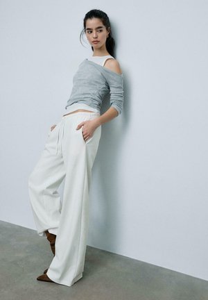 Mladá žena s tmavými vlasy sepnutými do culíku má na sobě šedý svetr se spadlými rameny, bílé kalhoty se širokými nohavicemi a hnědé špičaté boty, opírá se o zeď.