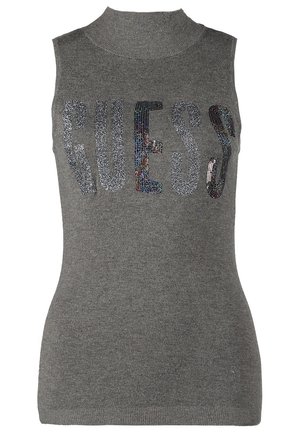 Vrouw met een grijze mouwloze coltrui met de tekst "GUESS", crème skinny broek, beige enkellaarsjes en een beige schoudertas met franjes.