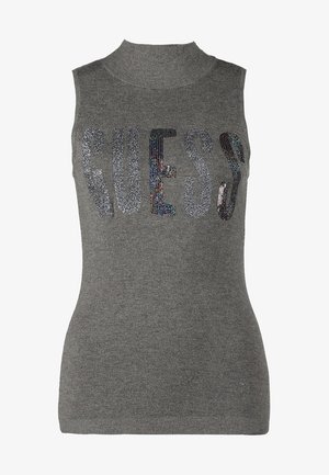 Frau trägt ein graues ärmelloses Rollkragenoberteil mit dem Schriftzug "GUESS", cremefarbene enge Hosen, sandfarbene Stiefeletten und trägt eine sandfarbene Schultertasche mit Fransen.