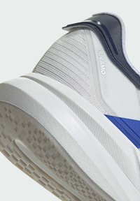 Chaussure de sport blanche avec accent bleu, tige texturée et semelle en caoutchouc. Présente un design profil bas avec un matériau durable et une traction à motif.
