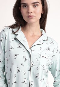 Camisa de pijama azul claro con un patrón divertido de personajes de dibujos animados. Tiene un ribete oscuro y un bolsillo en el lado izquierdo del pecho. Textura de tela suave.