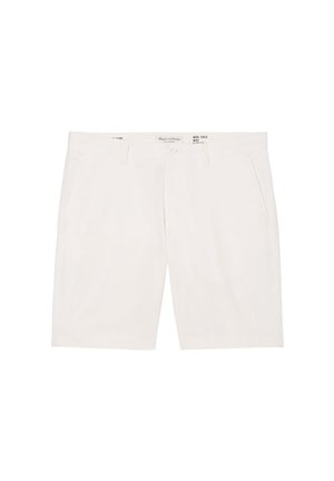 Personne portant une chemise à manches courtes violet clair, un short blanc à hauteur des genoux et des sandales beiges, debout contre un mur blanc uni.
