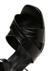 Sandal de tacón negro con un diseño entrecruzado y textura de cocodrilo. Suela lisa y punta abierta, con una silueta elegante.