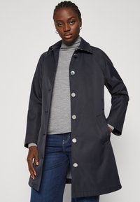 Cappotto impermeabile blu scuro con colletto, cinque bottoni metallici, tasche laterali e un tessuto liscio e impermeabile. Indossato sopra un dolcevita grigio.