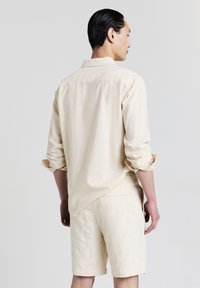 Uomo con capelli pettinati all'indietro, in piedi di spalle, indossa una camicia a maniche lunghe di tessuto color crema con i polsini arrotolati e pantaloni corti coordinati.