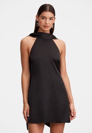 Femme aux cheveux raides et foncés portant une robe noire sans manches à col halter et des boucles d'oreilles géométriques rectangulaires sur un fond clair.