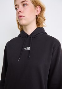 Črna kapuca iz mehke tkanine z srebrnim logojem "The North Face" na levem prsnem delu. Na voljo so strukturalne vrvice.