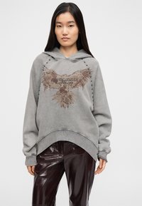 Γκρι φαρδιά μπλούζα με κουκούλα, με μπροστινό γραφικό αετού και το κείμενο "JUST CAVALLI". Διαθέτει αντιθετική ραφή και καμπυλωτή κάτω ρίγα, συνδυασμένη με λαμπερό μπορντό παντελόνι.