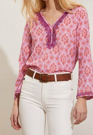 Femme portant un chemisier rose à motifs floraux avec une broderie violette et un pantalon blanc, accessoirisée d'une ceinture en cuir marron avec des œillets métalliques.