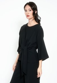 Top in tessuto nero con maniche ampie e vita legata. Presenta una scollatura rotonda e una texture liscia, creando una silhouette strutturata.