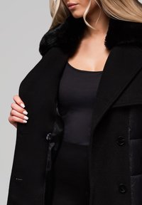 Kvinde, der viser en sort frakke med pelskrave over en sort top, mens hun holder frakken åben for at afsløre det indvendige foer.