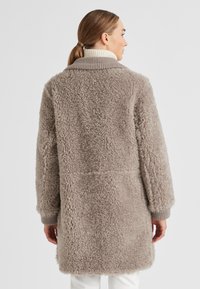 Frau mit leicht braunem Pferdeschwanz, die einen langen, plüschigen taupefarbenen Sherpa-Mantel und einen cremefarbenen gerippten Rollkragenpullover trägt, schaut von einer einfarbigen weißen Hintergrund weg.