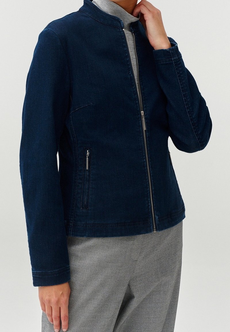 Person in einer dunkelblauen Zip-Jacke mit zwei seitlichen Reißverschlusstaschen und hellgrauen Hosen, Hand in der Nähe des Kragens.