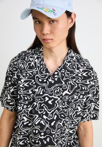 Camicia corta a maniche corte a motivo bianco e nero con forme astratte; cappellino azzurro con ricami floreali; collana d'argento.