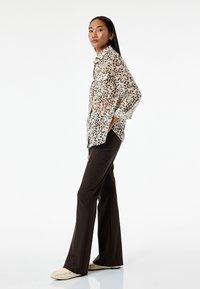 Transparente Bluse mit Tiermotiven in Braunflecken auf hellem Hintergrund, kombiniert mit dunkelbraunen Hosen und beigen Ballerinas, seitlich stehend.