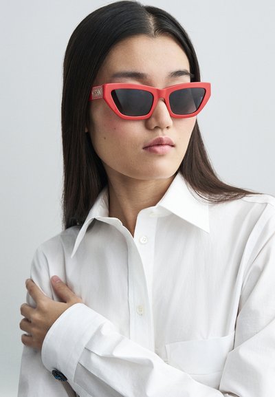 Punaiset kissa silmälasit tummilla linsseillä. Kumipintainen rakenne. Kultainen logo-aksentti varressa. Mallilla on päällään valkoinen napillinen paita.