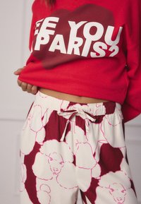 Hanorac roșu cu text alb "SEE YOU PARIS?", asociat cu pantaloni jogging crema cu ilustrații de pudel roz și talie cu șnur.