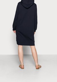 Navy blauwe hoodie jurk met lange mouwen, zijsplitten en geribbelde zoom. Draagt met bruine sandalen, wat een ontspannen silhouet toont.