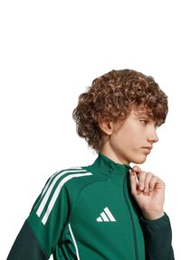 Groene sportjack met witte accenten en drie strepen op de schouders; zachte stof, ritskraag en logo op de borst.