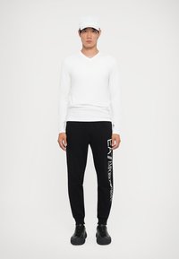 Cămașă albă cu mâneci lungi și decolteu în V, pantaloni de trening negri cu logo alb, adidași negri și o șapcă albă. Modelul stă drept în fața unui fundal simplu.
