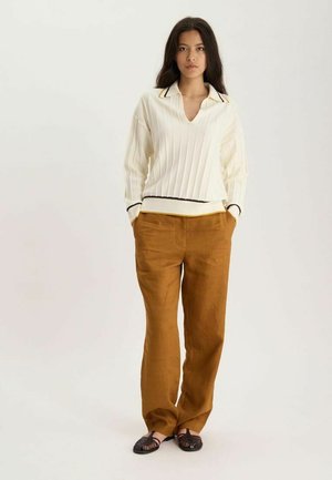Femme debout, les mains dans les poches, portant un pull blanc côtelé avec col, un pantalon marron à jambes larges et des ballerines tissées foncées sur fond uni.