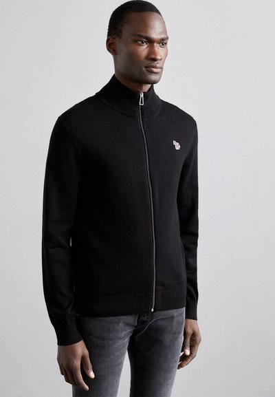 Cardigan zippé noir avec un col montant, texture lisse et logo discret sur la poitrine gauche. Associé à un jean foncé.