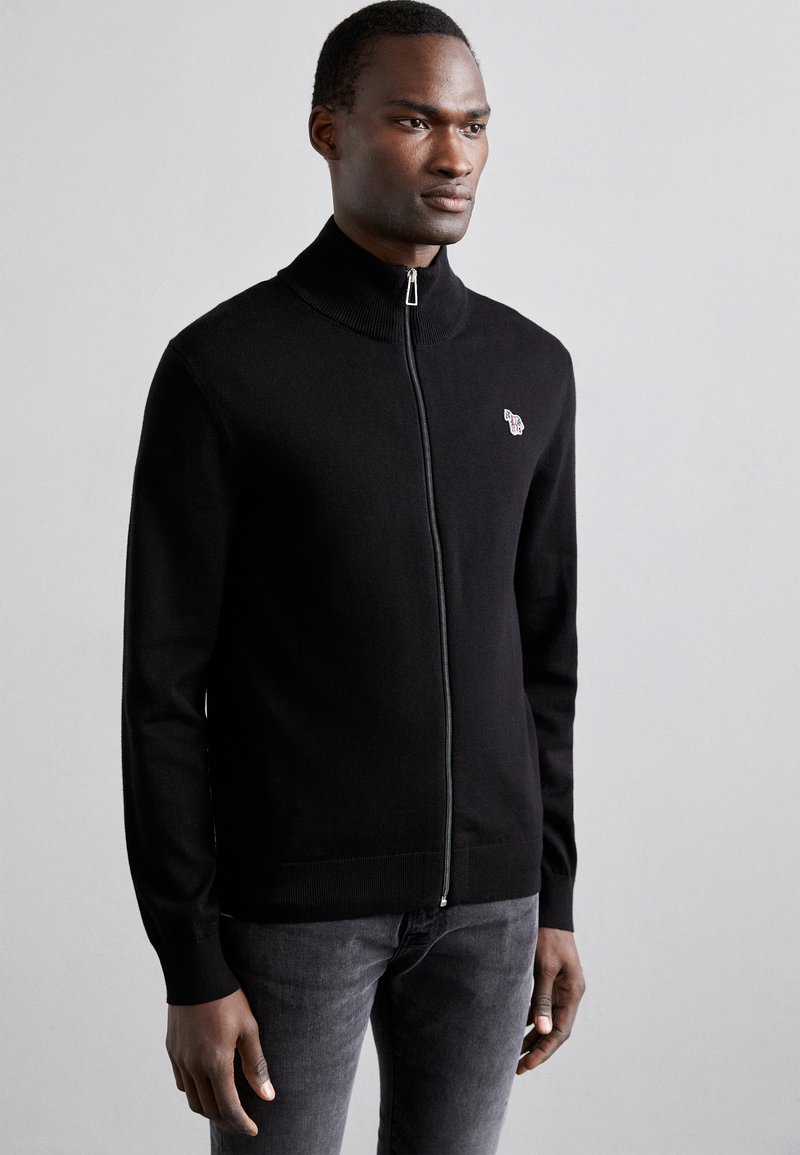 Black zip-up cardigan with a high collar, smooth texture, and subtle logo detail on the left chest. Paired with dark jeans.