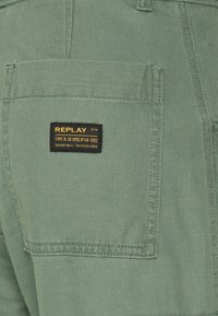 Calças de algodão verdes com um bolso traseiro onde se lê uma etiqueta preta "REPLAY". A costura é visível ao longo do bolso e das costuras.