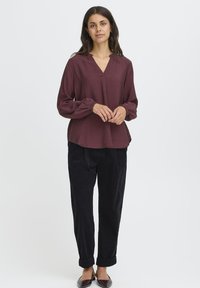 Femme debout portant un blouse bordeaux à manches longues, un pantalon noir retroussé et des ballerines noires à bout pointu, le tout sur un fond clair et uni.