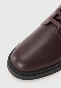 Scarpa in pelle marrone scuro con finitura lucida, punta arrotondata e suola nera texturizzata. Presenta lacci beige e una superficie superiore liscia.