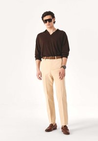 Ruda ilgomis rankovėmis polo marškinėliai, smėlio spalvos siūtos kelnės, rudi odiniai batai, rudas diržas ir tamsūs akiniai nuo saulės ant vienspalvio fono.