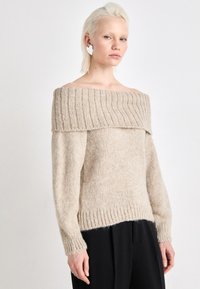 Beige off-shoulder tröja med ribbad halsringning och ärmslut, texturerad stickad tyg och lös passform, matchad med svarta byxor.