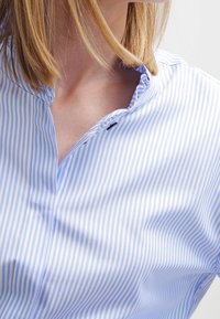 Niebieska i biała koszula z kołnierzykiem w paski, z zapięciem na guziki i miękką teksturą. Charakteryzuje się wąskimi pionowymi paskami o gładkim wykończeniu.