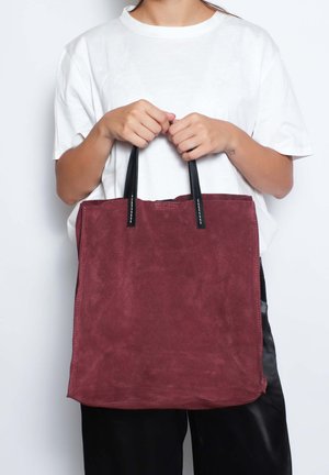 Una persona con una camicia bianca tiene davanti a uno sfondo bianco una grande borsa tote in suede marrone scuro con manici neri.