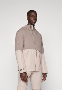 Lightweight, two-tone jacket in beige and taupe, featuring a high collar, zip closure, and zippered pockets. Made of smooth fabric.